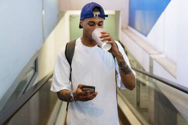 Tattooed young man with backpack standing on escalator looking at cell phone while drinking coffee to go - MGIF00542