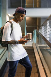 Young man with backpack and coffee to go standing on escalator looking at cell phone - MGIF00543