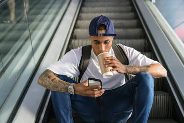 Junger Mann mit Rucksack, der auf einer Rolltreppe sitzt und auf sein Handy schaut, während er einen Coffee to go trinkt - MGIF00544