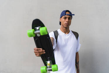 Porträt eines tätowierten jungen Mannes mit Skateboard vor einem grauen Hintergrund - MGIF00558