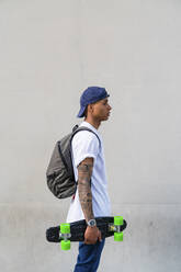 Tattooed young man with skateboard and backpack in front of grey background - MGIF00559