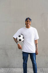 Portrait of tattooed young man with football in front of concrete wall - MGIF00561