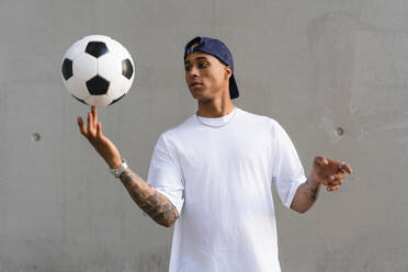 Portrait of tattooed young man balancing football on his finger in front of concrete wall - MGIF00565