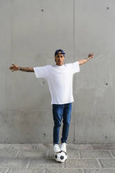 Portrait of tattooed young man balancing on football in front of concrete wall - MGIF00568