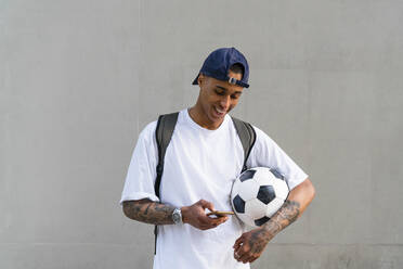 Tätowierter junger Mann mit Fußball, der auf sein Smartphone schaut - MGIF00569