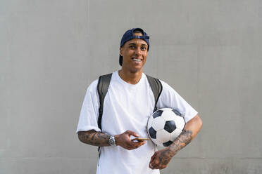 Portrait of tattooed young man with football and smartphone in front of concrete wall - MGIF00570