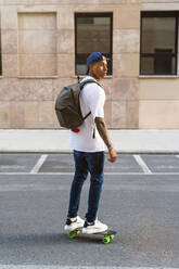 Tattooed young man with backpack standing on skateboard looking around - MGIF00573