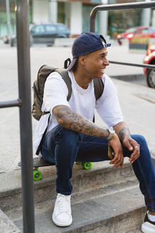 Tätowierter junger Mann mit Rucksack und Smartphone sitzt auf einem Skateboard und schaut in die Ferne - MGIF00577