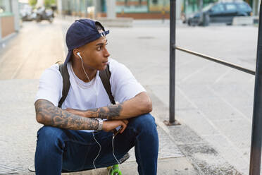 Tattooed young man sitting on his skateboard listening music with smartphone and earphones - MGIF00580