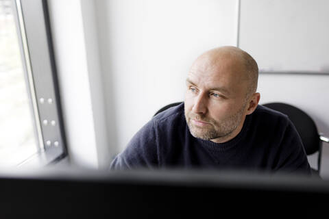 Nachdenklicher Mann, der vor einem Computer sitzt und aus dem Fenster schaut, lizenzfreies Stockfoto
