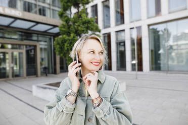 Lächelnde Frau in der Stadt, die draußen steht und ihr Smartphone benutzt - KMKF01013