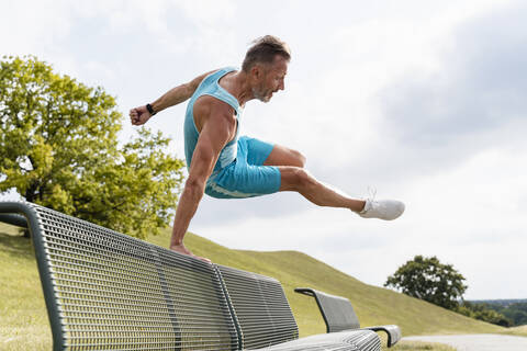 Sportlicher Mann springt über eine Bank im Park, lizenzfreies Stockfoto