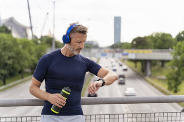 Sporty man standing on a bridge, wearing headphones and checking his smartwatch - DIGF07554