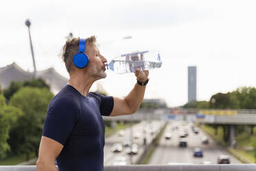 Sportlicher Mann mit Kopfhörern, der auf einer Brücke Wasser trinkt - DIGF07559