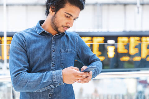 Junger Mann mit Jeanshemd und Smartphone am Bahnhof, London, Großbritannien, lizenzfreies Stockfoto