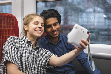 Porträt eines glücklichen jungen Paares, das ein Selfie mit einer Sofortbildkamera im Zug macht, London, UK - WPEF01597