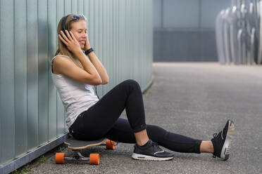 Smiling young woman sitting on longboard listening music with headphones - STSF02122