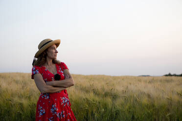 Frau mit Strohhut und rotem Sommerkleid mit Blumenmuster steht vor einem Getreidefeld - FLLF00242