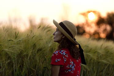 Woman in front of grain field wearing straw hat and red summer dress with floral design at sunset - FLLF00246