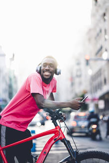 Glücklicher junger Mann mit E-Bike und Smartphone in der Stadt - OCMF00497