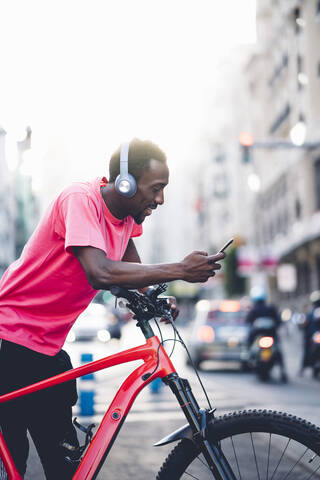 Junger Mann mit E-Bike und Kopfhörern, der in der Stadt sein Smartphone benutzt, lizenzfreies Stockfoto