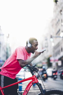 Junger Mann mit E-Bike und Kopfhörern, der in der Stadt sein Smartphone benutzt - OCMF00499