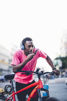 Junger Mann mit E-Bike und Kopfhörern, der in der Stadt sein Smartphone benutzt - OCMF00500