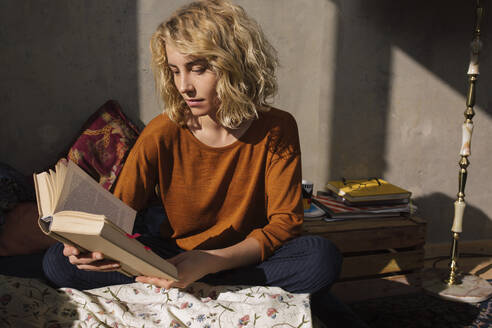 Blond student sitting on bed reading a book - GCF00256