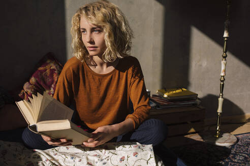 Porträt einer nachdenklichen blonden Studentin, die mit einem Buch auf dem Bett sitzt und in die Ferne schaut - GCF00257