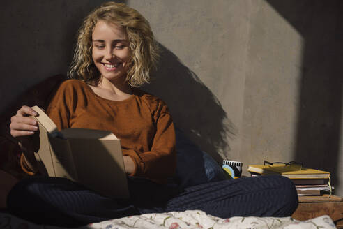 Porträt einer lächelnden jungen Frau, die auf einem Bett sitzt und ein Buch liest - GCF00258