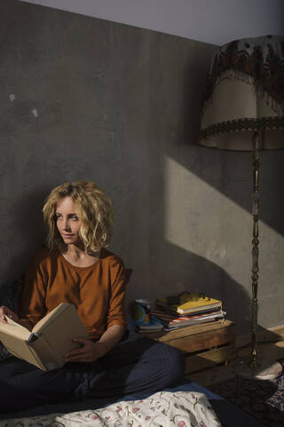 Blonde Studentin sitzt mit einem Buch auf dem Bett und schaut in die Ferne, lizenzfreies Stockfoto