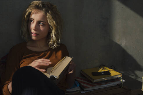 Blond student sitting on bed with a book looking at distance - GCF00268