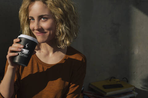 Porträt einer lächelnden jungen Frau, die zu Hause Kaffee zum Mitnehmen trinkt, lizenzfreies Stockfoto