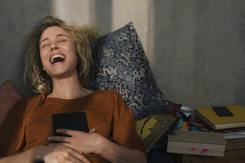 Portrait of laughing young woman lying on bed with E-book reader - GCF00279