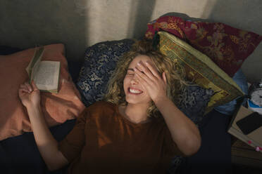 Portrait of laughing young woman lying on bed reading a book - GCF00283