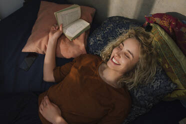 Portrait of laughing young woman with eyes closed lying on bed with a book - GCF00285