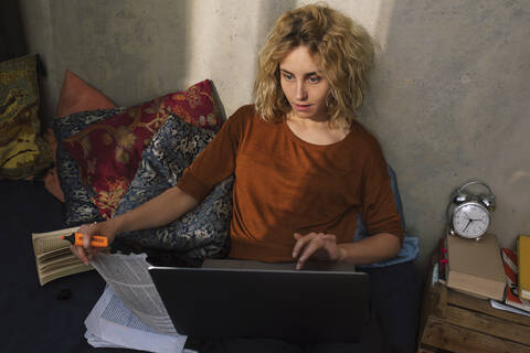 Porträt einer blonden Studentin, die auf einem Bett sitzt und an einem Laptop arbeitet, lizenzfreies Stockfoto