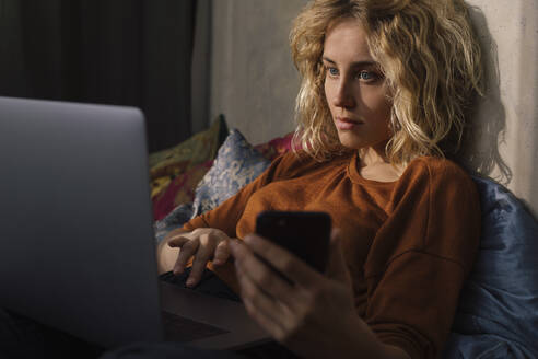 Portrait of blond young woman lying on bed using cell phone and laptop - GCF00290