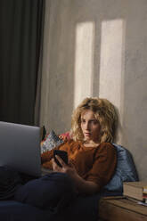 Porträt einer blonden jungen Frau auf dem Bett liegend mit Handy und Laptop - GCF00292