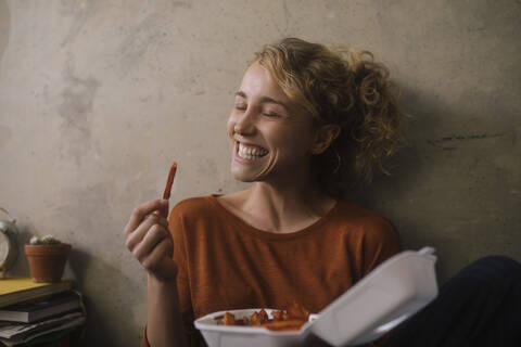 Porträt einer grinsenden jungen Frau, die zu Hause Pommes frites isst, lizenzfreies Stockfoto