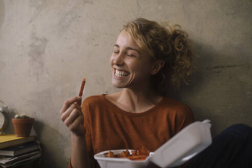 Porträt einer grinsenden jungen Frau, die zu Hause Pommes frites isst - GCF00309