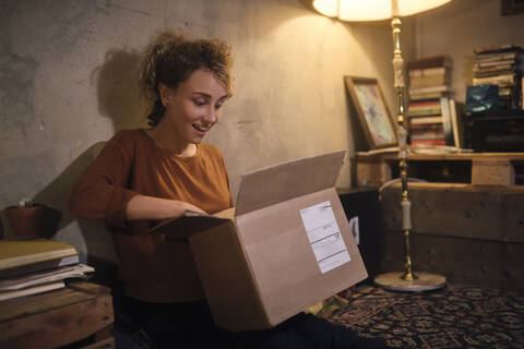 Erstaunte junge Frau beim Auspacken eines Pakets zu Hause, lizenzfreies Stockfoto