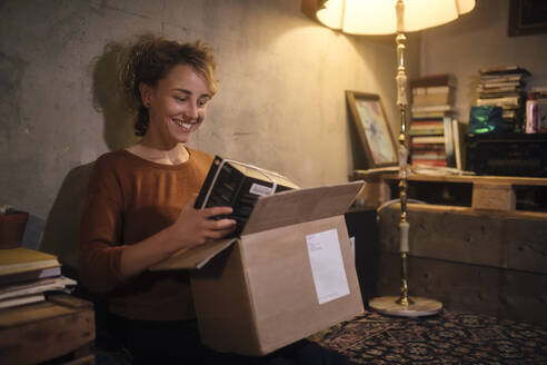 Lächelnde junge Frau beim Auspacken eines Pakets zu Hause - GCF00315