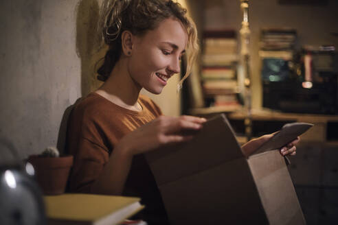 Lächelnde junge Frau beim Auspacken eines Pakets zu Hause - GCF00316