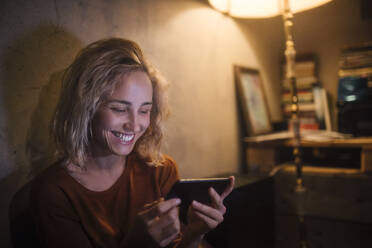 Porträt einer lachenden jungen Frau, die zu Hause ihr Smartphone benutzt - GCF00322