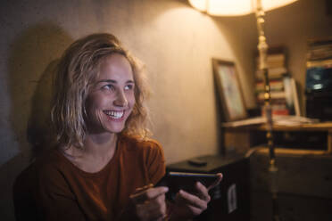 Porträt einer lachenden jungen Frau, die zu Hause ihr Smartphone benutzt - GCF00323