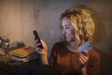 Blonde junge Frau benutzt Smartphone und Kreditkarte zu Hause - GCF00327