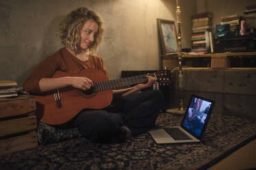 Lächelnde junge Frau sitzt mit Gitarre auf dem Boden und schaut auf einen Laptop - GCF00331