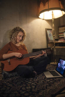 Junge Frau sitzt mit Gitarre auf dem Boden und schaut auf einen Laptop - GCF00332