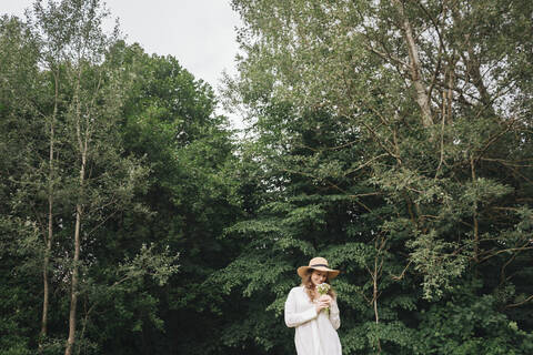 Junge Frau mit Hut hält einen Blumenstrauß vor einem Wald, lizenzfreies Stockfoto